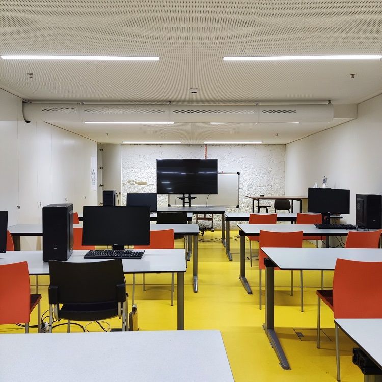 Computerlokaal met oranje stoelen, computers en een groot centraal scherm. Gele vloer, witte muren en witte geperforeerde metalen luchtverdeelkanalen.