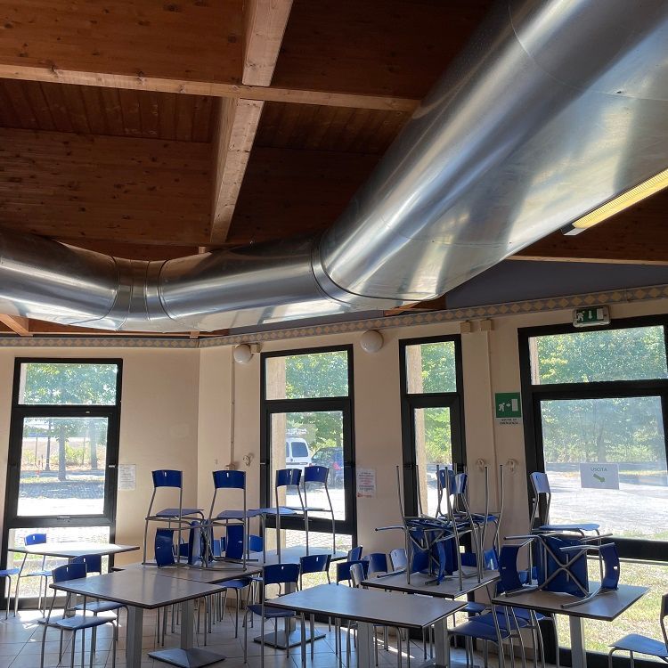 Cafetaria met vierkante tafels, opgestapelde blauwe stoelen, grote metalen luchtverdeelkanalen en ramen met uitzicht op groen.