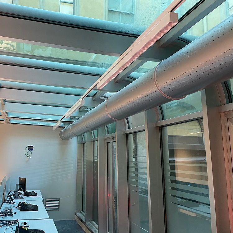 Interieur van een kamer met een glazen dak, een lange metalen ventilatiebuis, witte tafels, computers en geperforeerde metalen luchtverdeelkanalen.