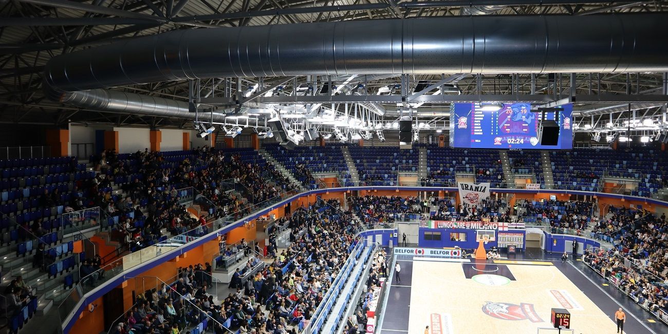 Basketbalarena volle tribunes. Spelers op het veld; scorebord zichtbaar. Blauwe stoelen met oranje accenten. met micro geperforeerde metalen luchtverdeelkanalen