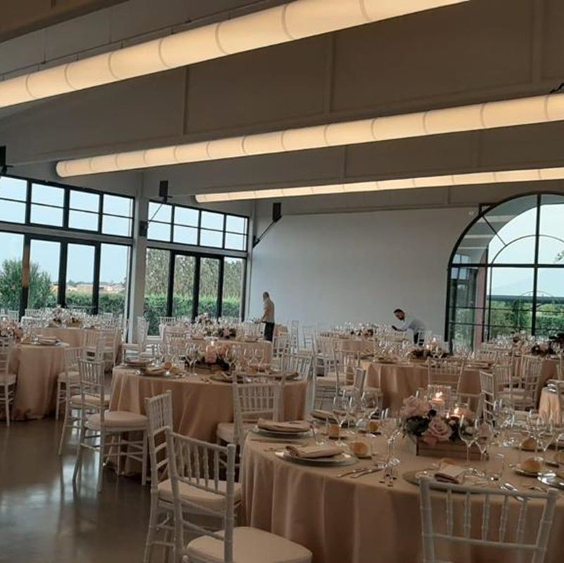 Elegante aankleding voor de huwelijksreceptie: ronde tafels met crèmekleurige tafelkleden, witte stoelen, bloemstukken en grote ramen voorzien van luchtverdeelkanalen & airsocks met geintegreerde verlichting