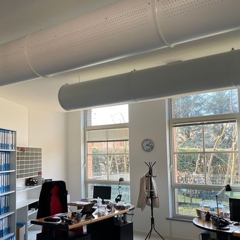 Kantoor met twee grote ramen, bureau, planken en geperforeerde metalen ventilatiekanalen.