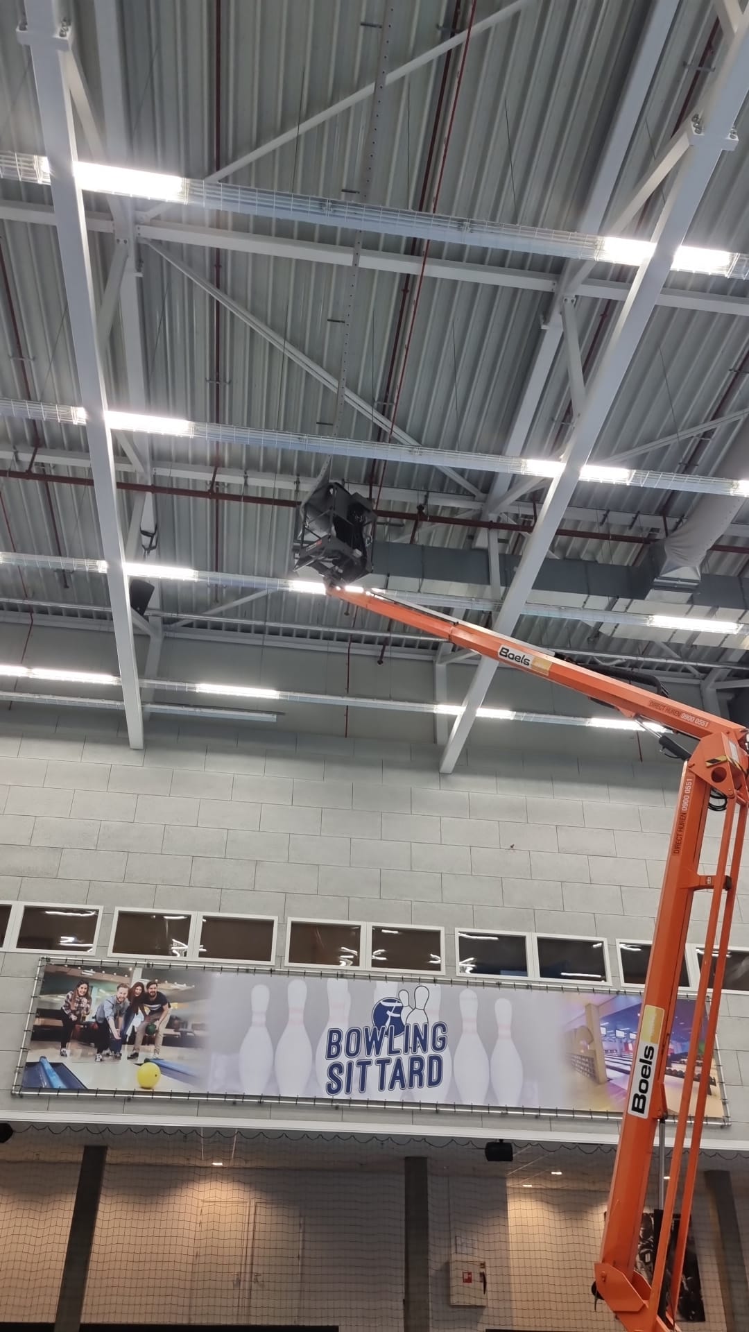 Een hoogwerker is aan het werk aan airsocks/luchtverdeelslangen aan het plafond van een gebouw.