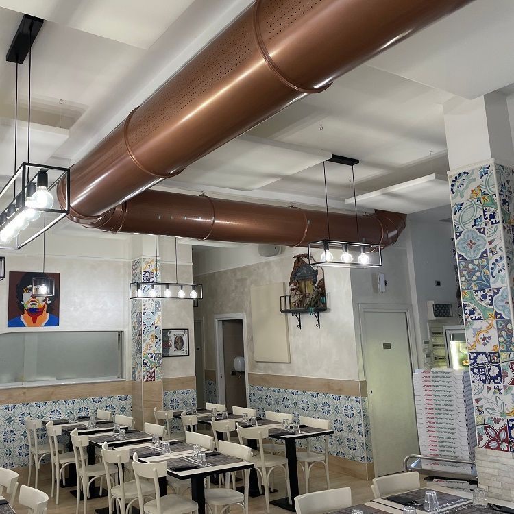Restaurantinterieur met koperkleurige geperforeerde metalen luchtkanalen, witte tafels en muren met decoratieve tegels.