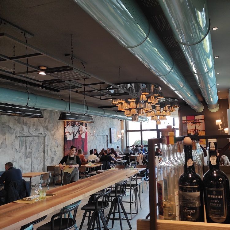 Restaurantinterieur: gasten aan tafels, een lange bar met barkrukken, metalen geperforeerde luchtverdeelkanalen en wijnflessen.