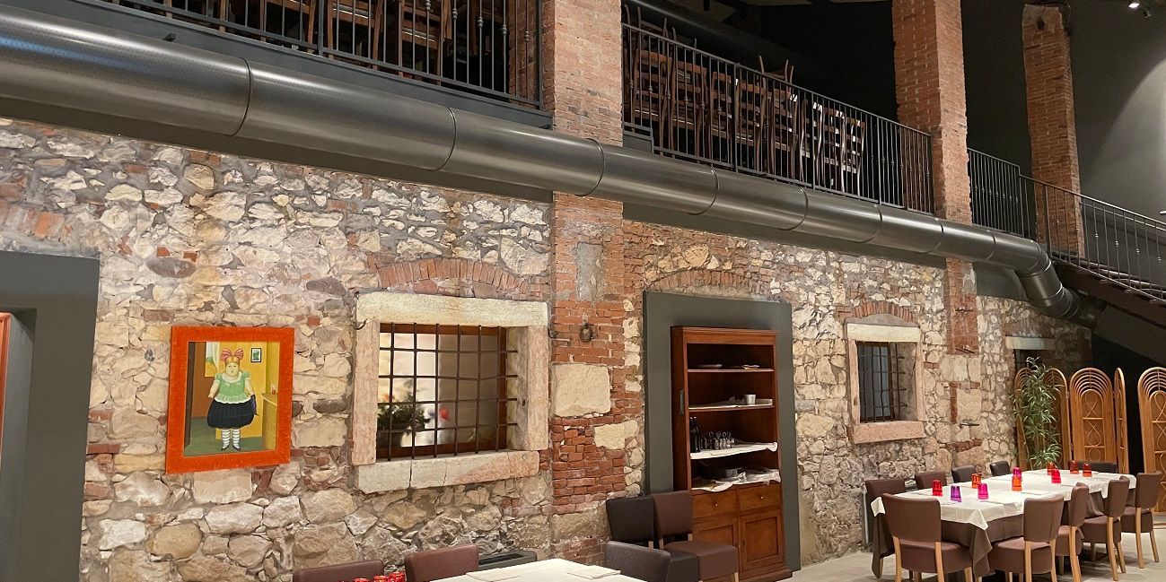 Een interieur met stenen muren, een eethoek, houten planken en een balkon met smeedijzeren balustrade en geperforeerde metalen luchtverdeelkanalen.