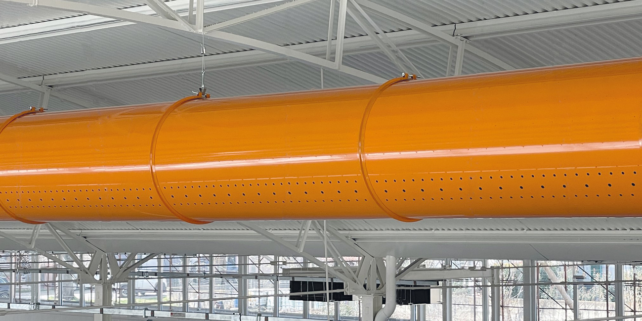 Oranje metalen geperforeerde luchtverdeelkanalen, opgehangen aan een wit plafond met steunbalken.