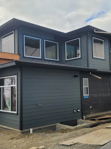Two-story house under construction with dark gray siding and multiple windows against a cloudy sky.
