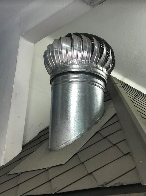 Ventilador de techo metálico con tapa de turbina plateada en un tejado de tejas.