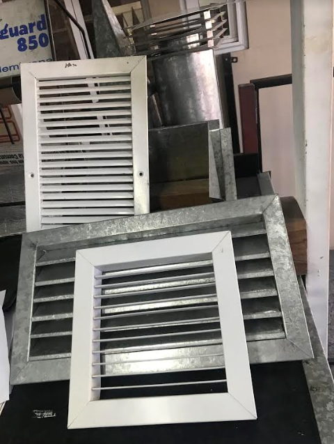 Rejillas de ventilación metálicas y paneles laminados apilados contra una pared en un taller.