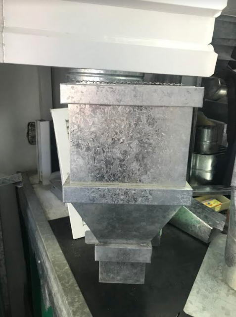 Tolva metálica para maquinaria industrial con embudo cuadrado y tapa con brida, mostrada en primer plano dentro de un taller.