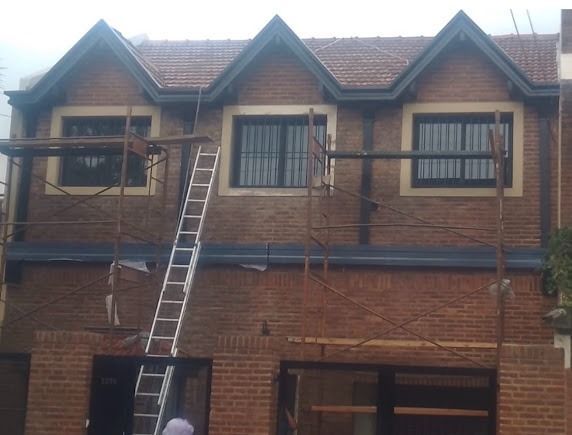 Escalera apoyada contra una casa de ladrillo con ventanas en la parte superior y tejado a dos aguas.