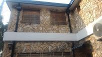 Fachada de edificio revestida de piedra con dos ventanas con contraventanas y una barandilla de balcón blanca.