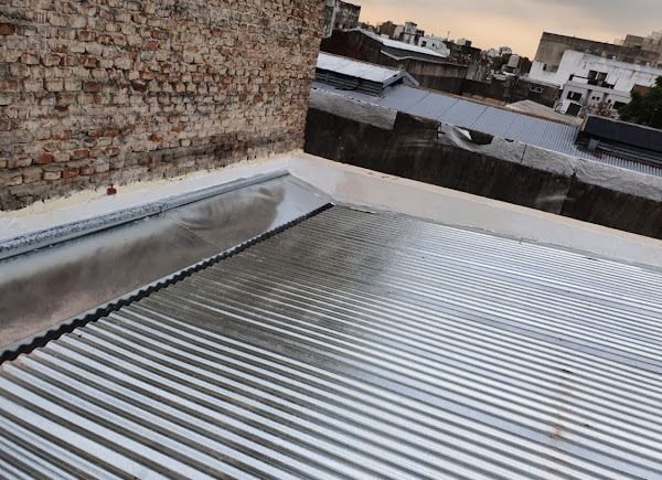 Azotea con techo de chapa ondulada junto a una pared de ladrillos y edificios vecinos al fondo.