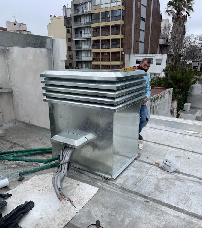 Unidad metálica de climatización en la azotea de un tejado plano, con un trabajador a su lado y edificios de la ciudad al fondo.