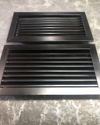 Dos rejillas de ventilación rectangulares negras sobre una superficie gris, una encima de la otra, mostradas en ángulo.