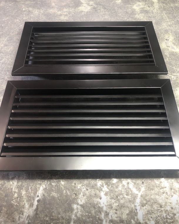 Dos rejillas de ventilación rectangulares negras con lamas sobre una superficie gris, una más pequeña encima de otra más grande.