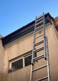 Una escalera apoyada contra una casa de color beige bajo un cielo azul, que llega hasta el tejado, justo encima de una ventana.