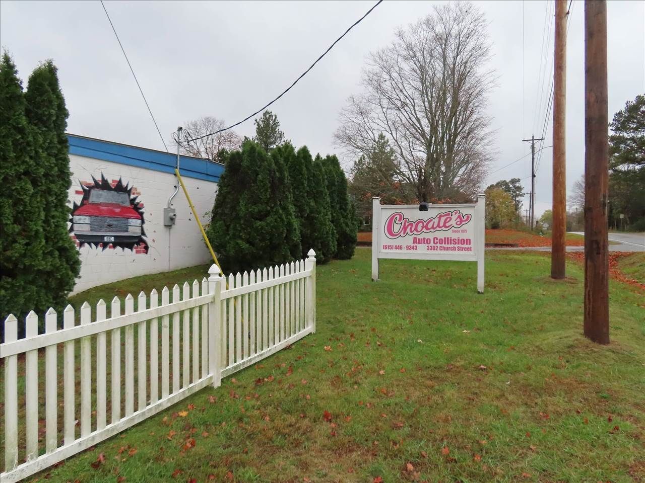Grassy roadside with a white fence, evergreen trees, and a red-and-white sign beside a building