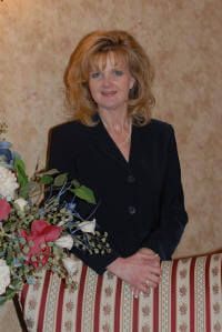 A woman in a black suit is sitting on a striped chair next to a vase of flowers.