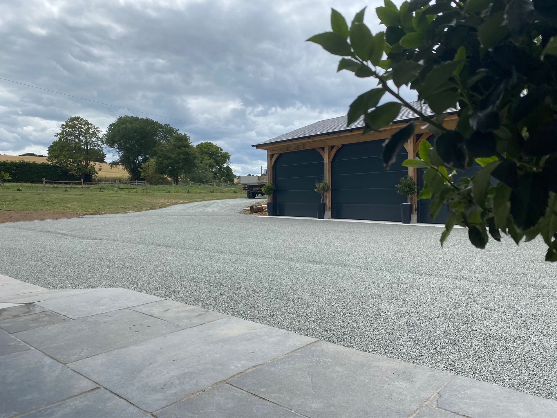 Gravel driveway leading to a two-bay garage with dark doors and a wooden frame under a cloudy sky.