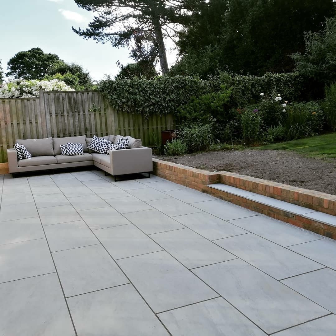 Patio with gray stone tiles and beige sectional sofa. Brick retaining wall and wooden fence in the background.
