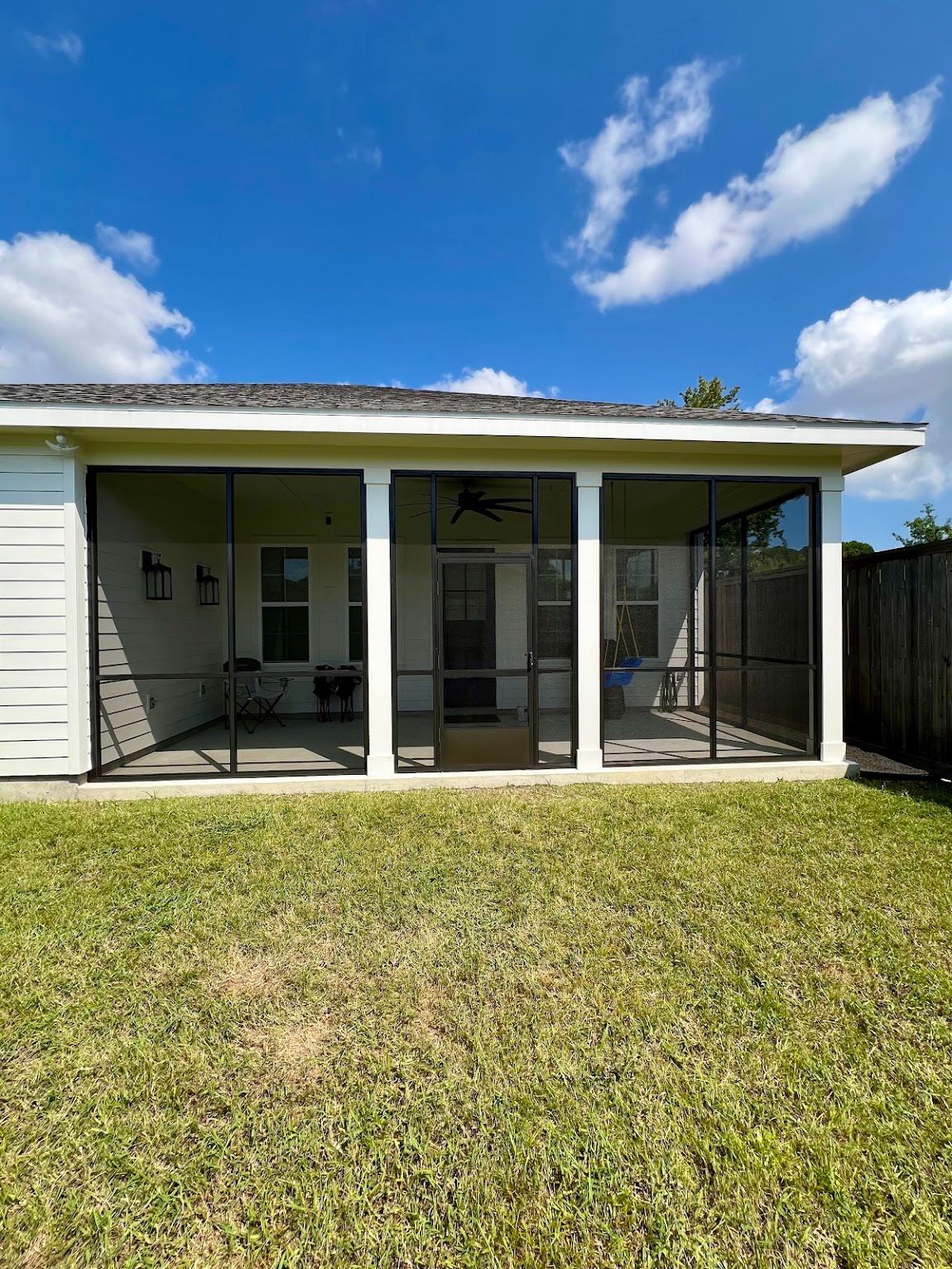 Covered backyard patio with screened enclosure beside a white house, under a blue sky.