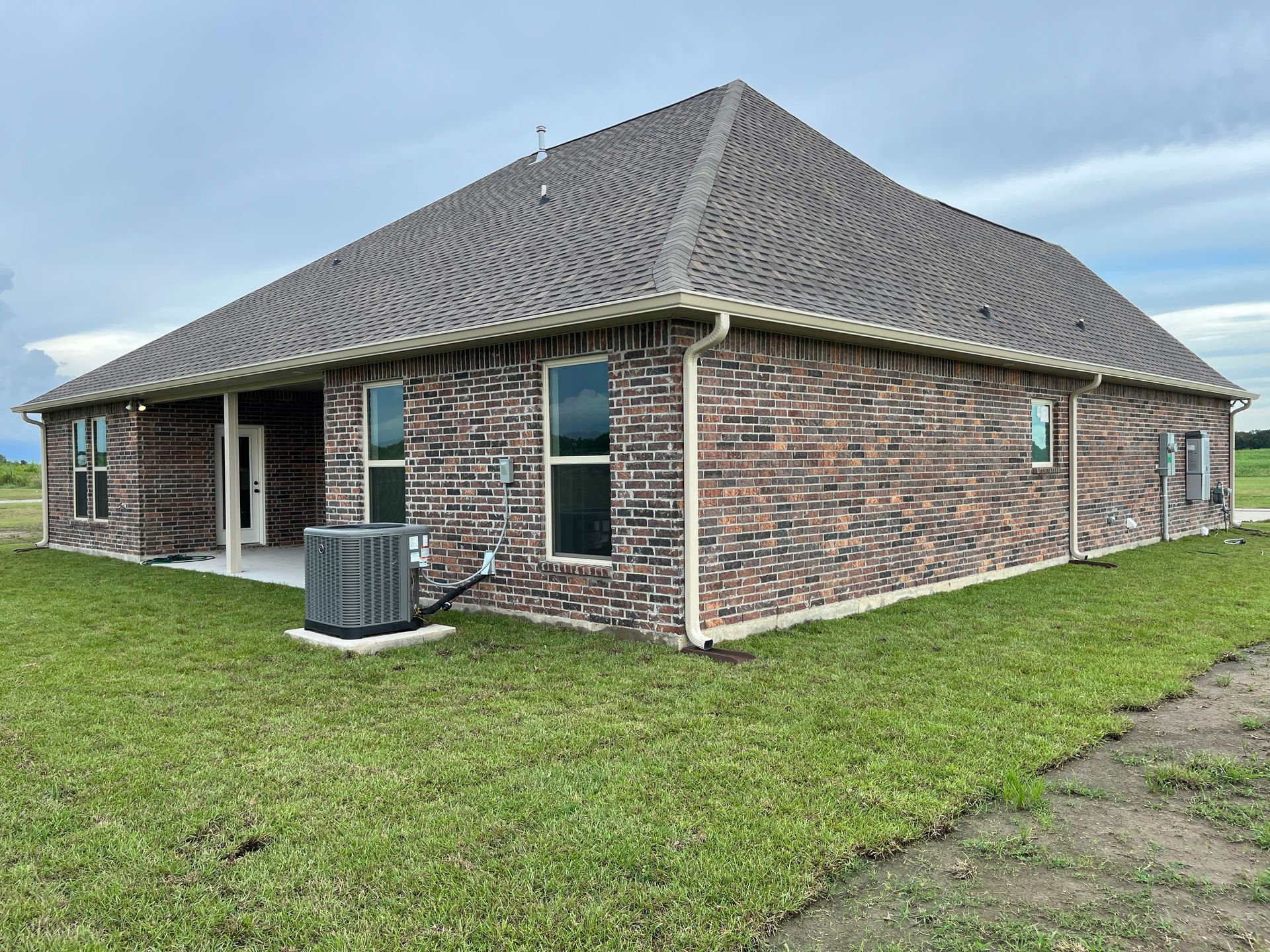 A single-story brick house with a dark shingle roof and an air conditioning unit on a grassy lawn under a cloudy sky.