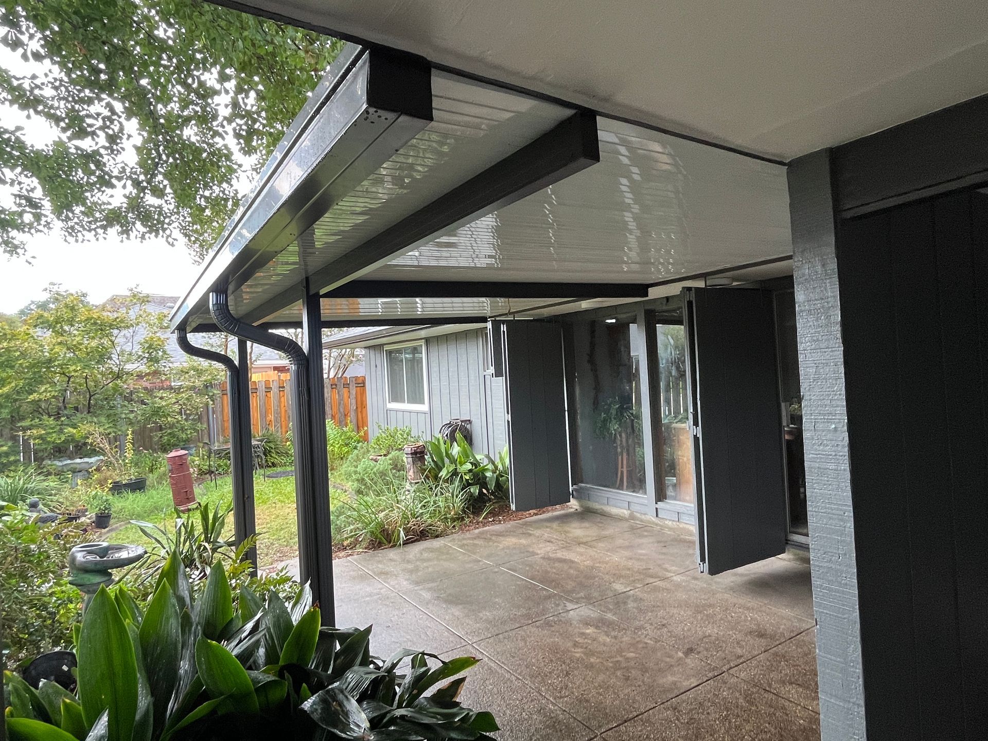 A covered patio with a white ceiling and black posts overlooking a garden on a rainy day.