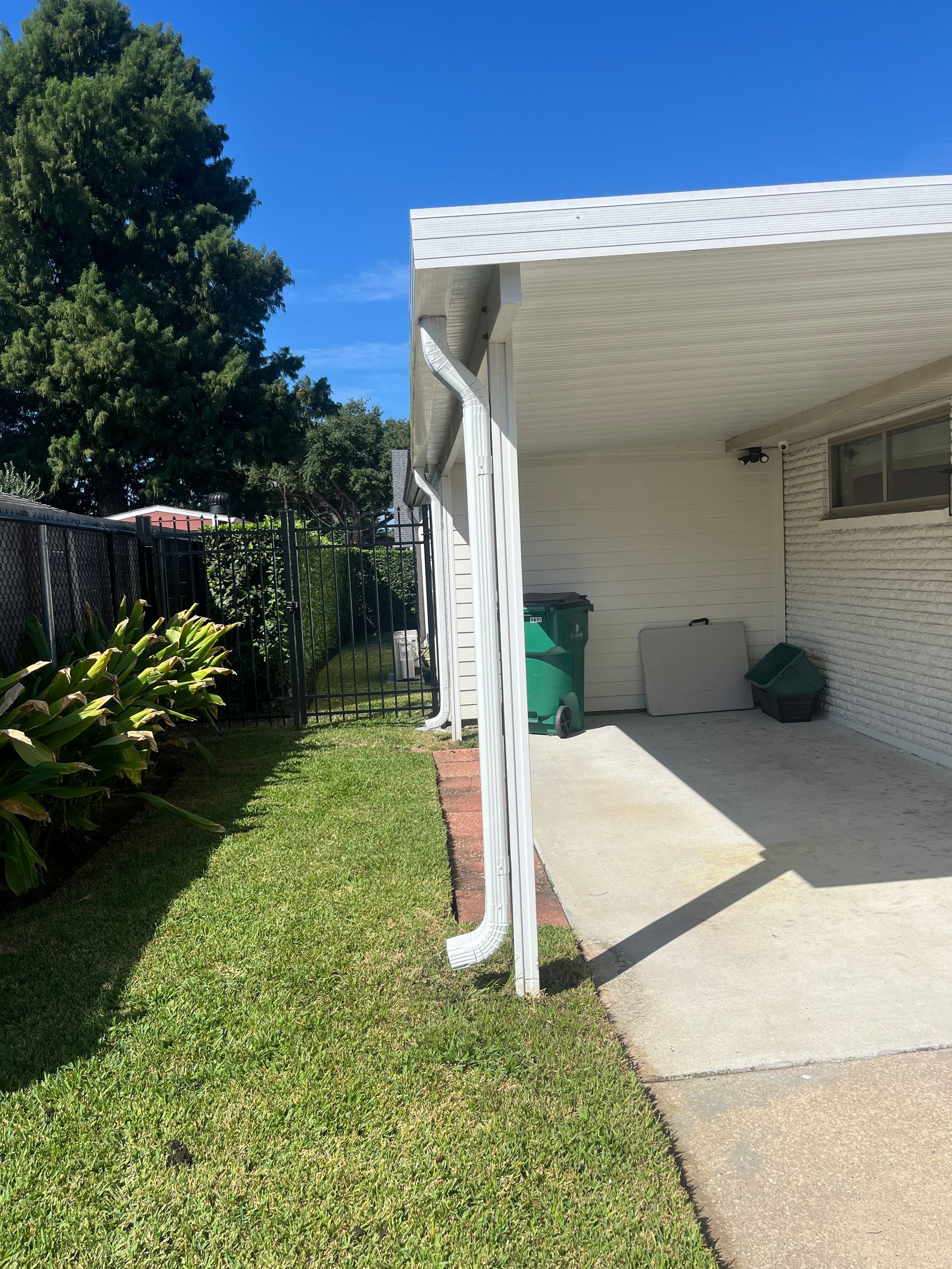 A white covered patio area with a concrete floor, a green trash bin, and a grassy lawn alongside a chain-link fence.