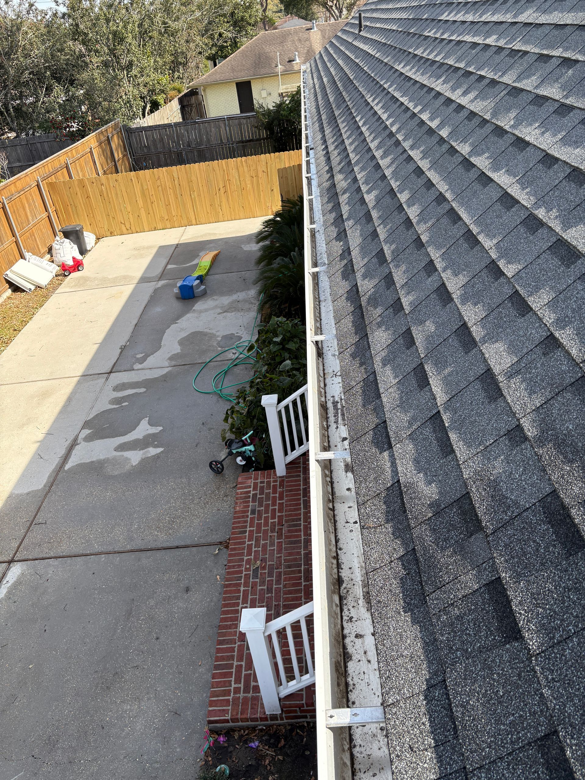 High-angle view of a roof edge with white gutters, overlooking a paved driveway, brick patio, and a wooden fence.