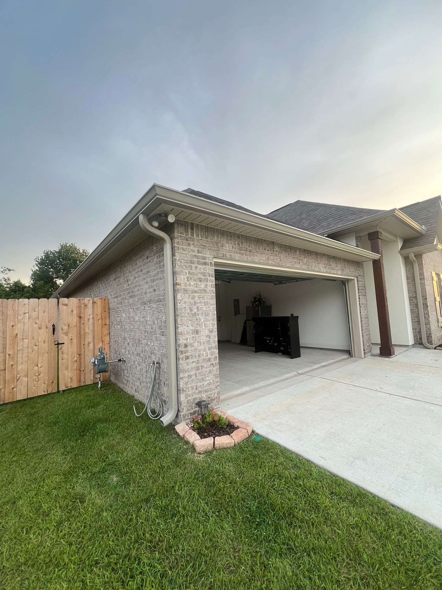 An open garage with light brick siding and a tan fence, situated next to a house under a cloudy sky.