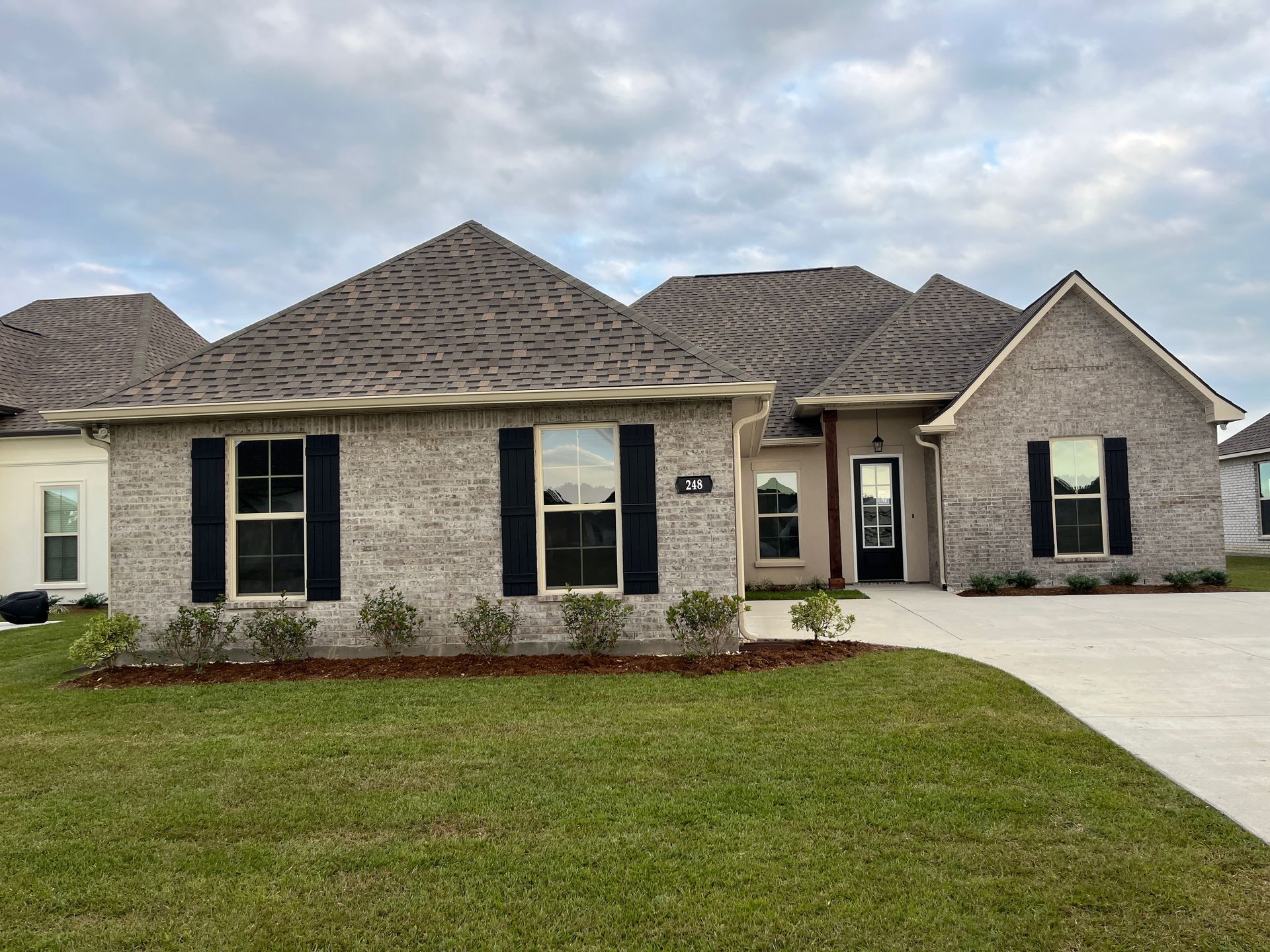 A single-story gray brick house with dark shutters, a front door, and a concrete driveway on a sunny day.