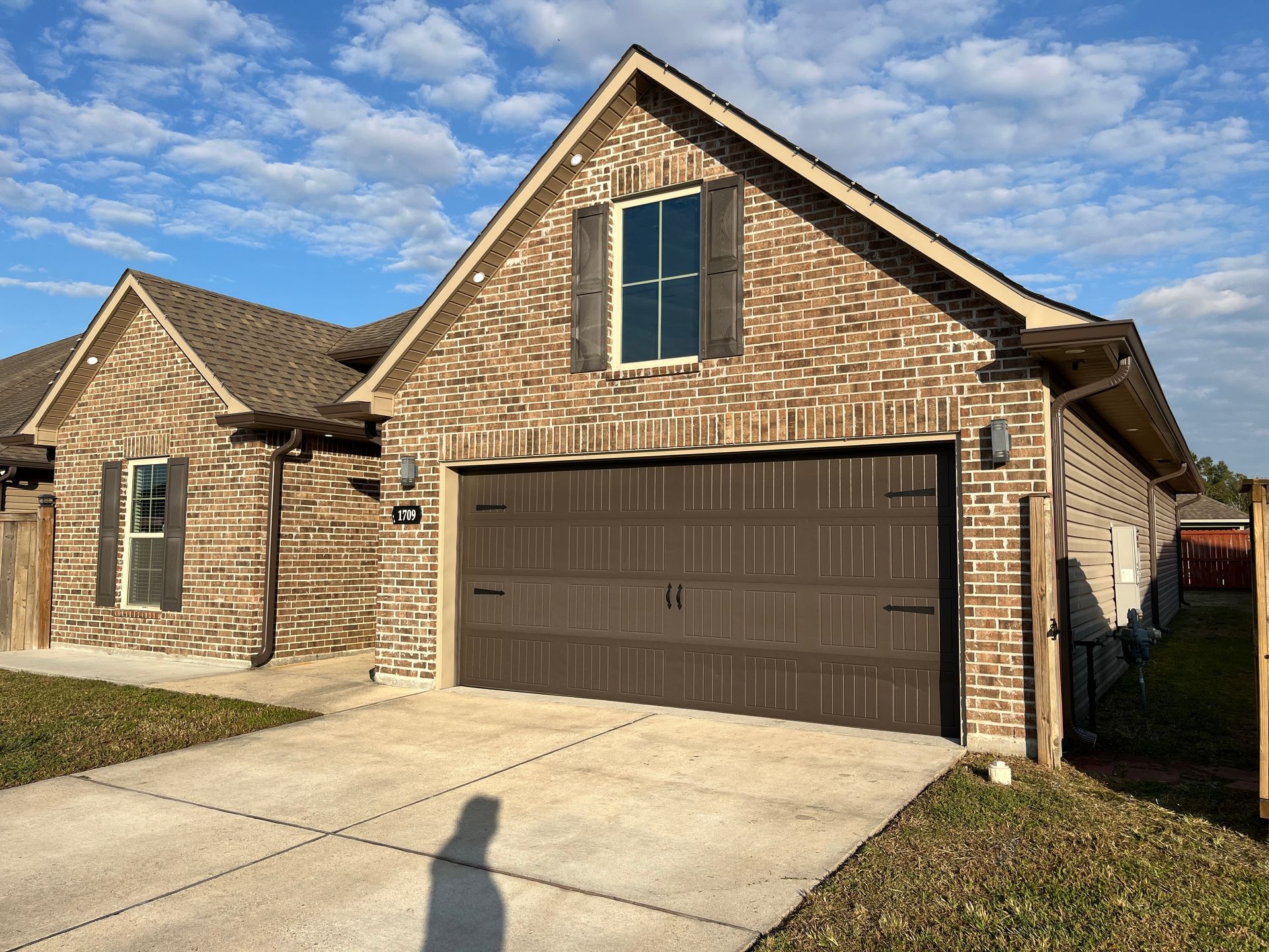 A brown brick house with a dark garage door, a concrete driveway, and shutters on the upper window under a blue sky.