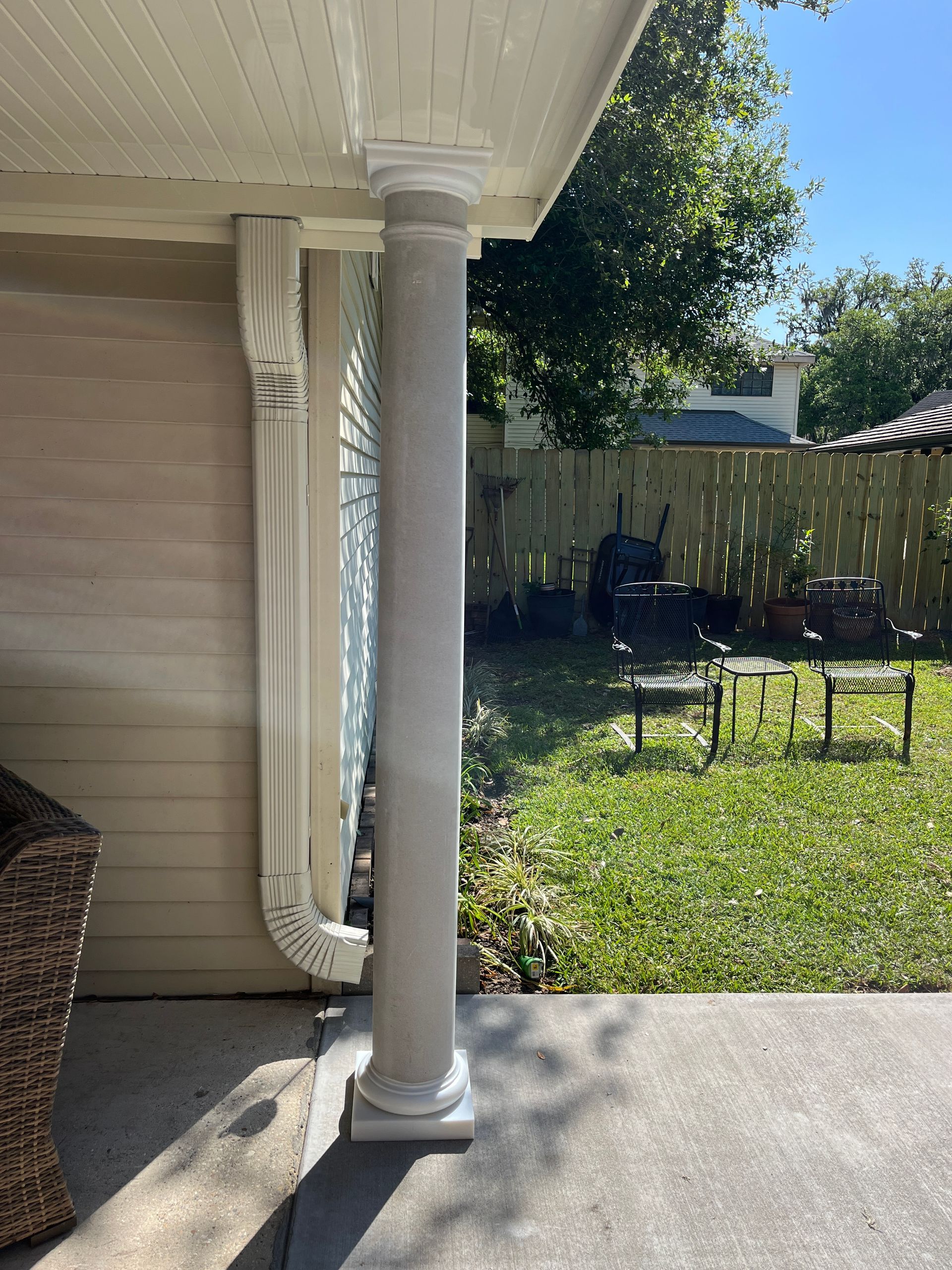A white porch column stands next to a downspout on a concrete patio, with a grassy yard and furniture in the background.