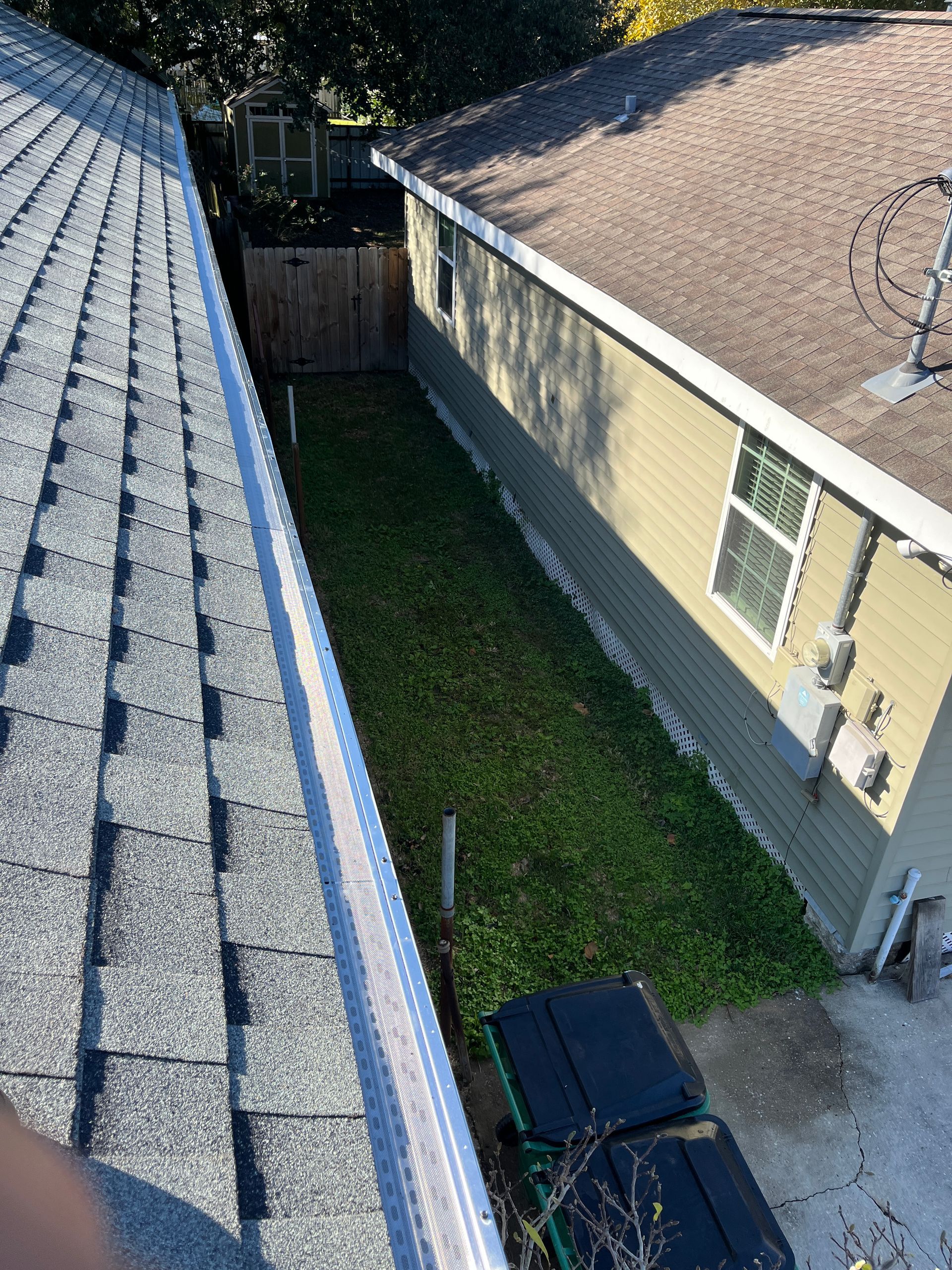 A high-angle view looking down at a narrow grassy strip between two houses with shingled roofs.
