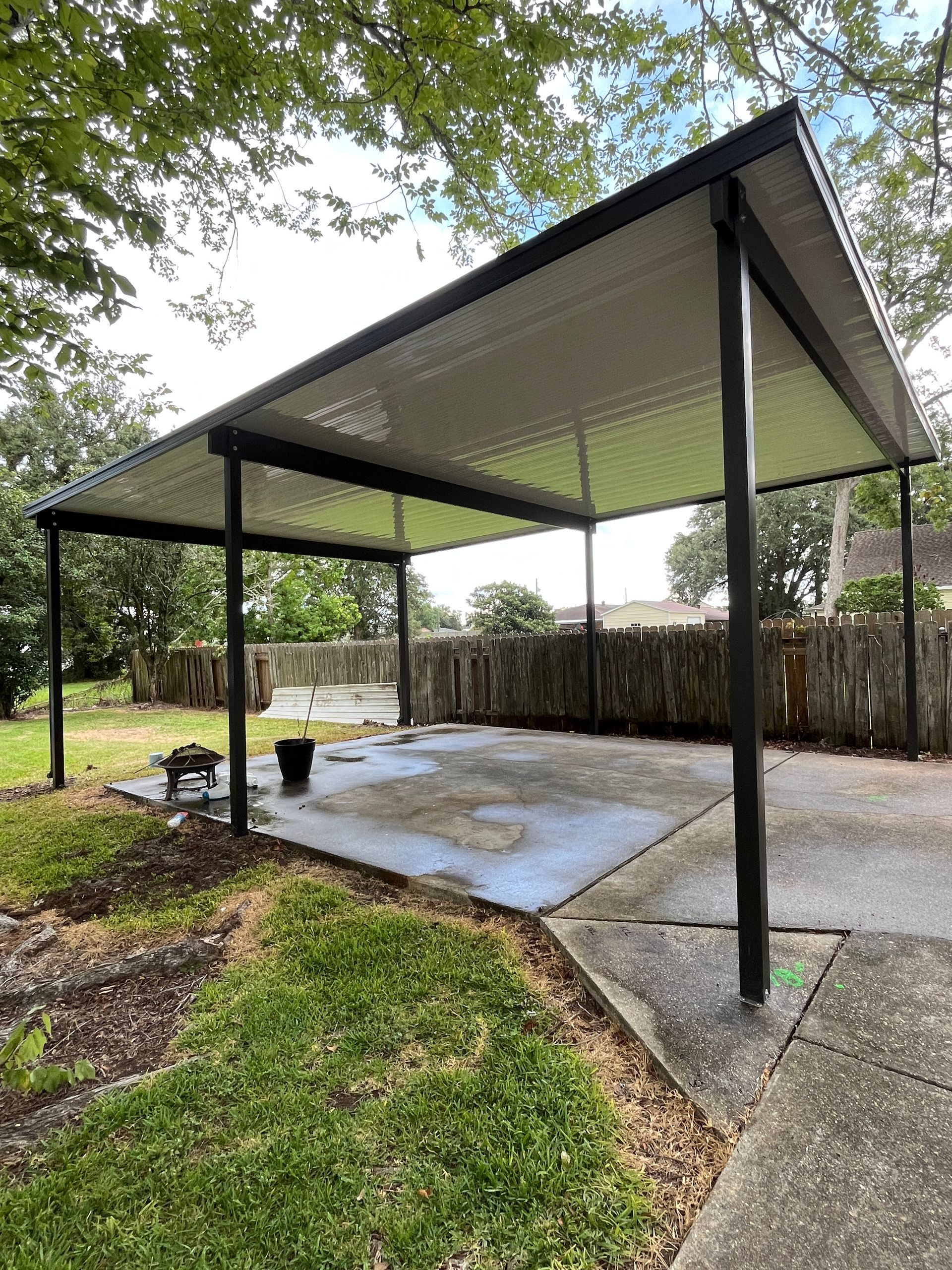 A modern black-framed patio cover with a white corrugated roof, situated over a concrete slab in a grassy backyard.