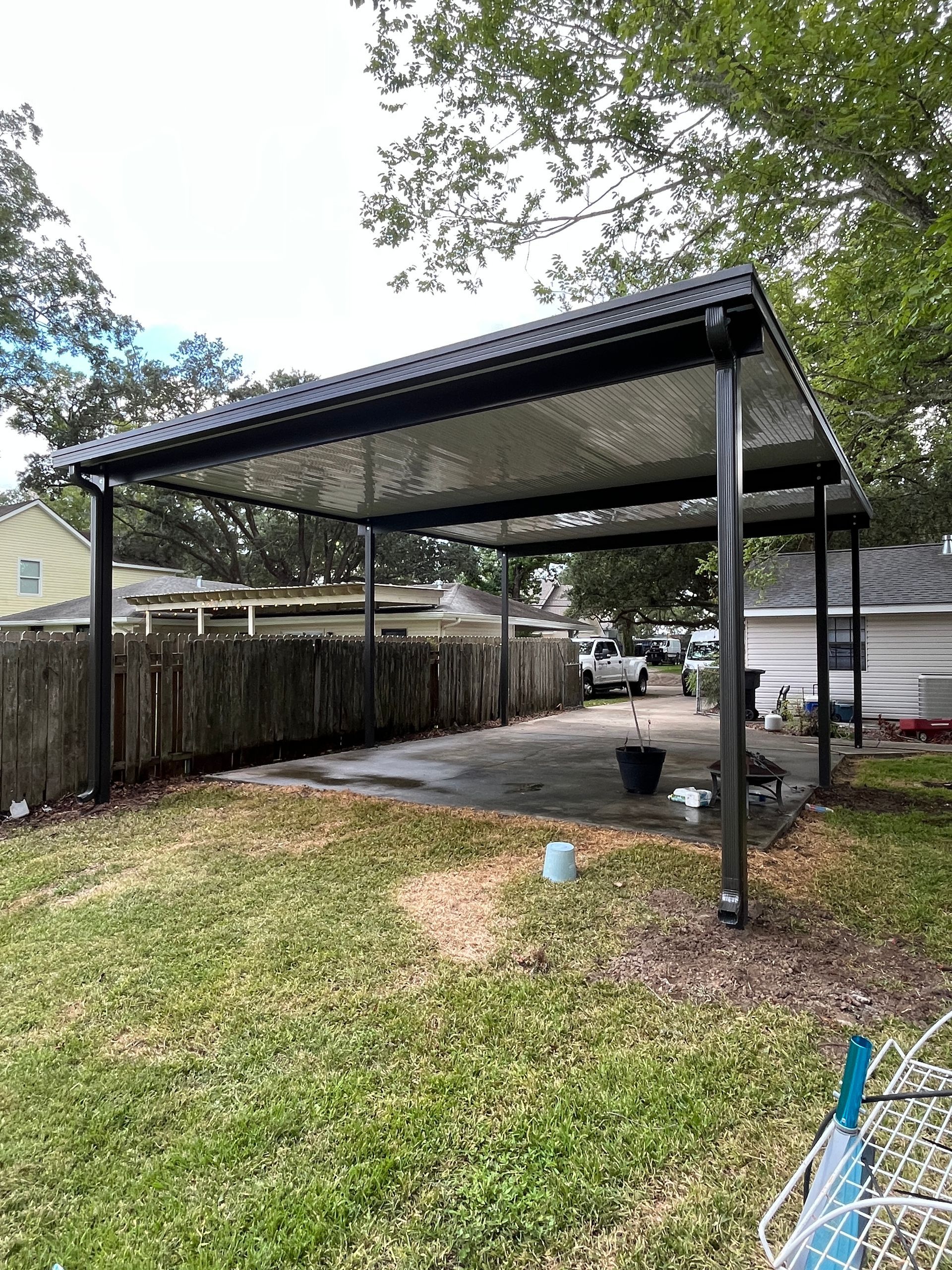 A black metal carport stands over a concrete driveway in a backyard with a wooden fence and surrounding trees.
