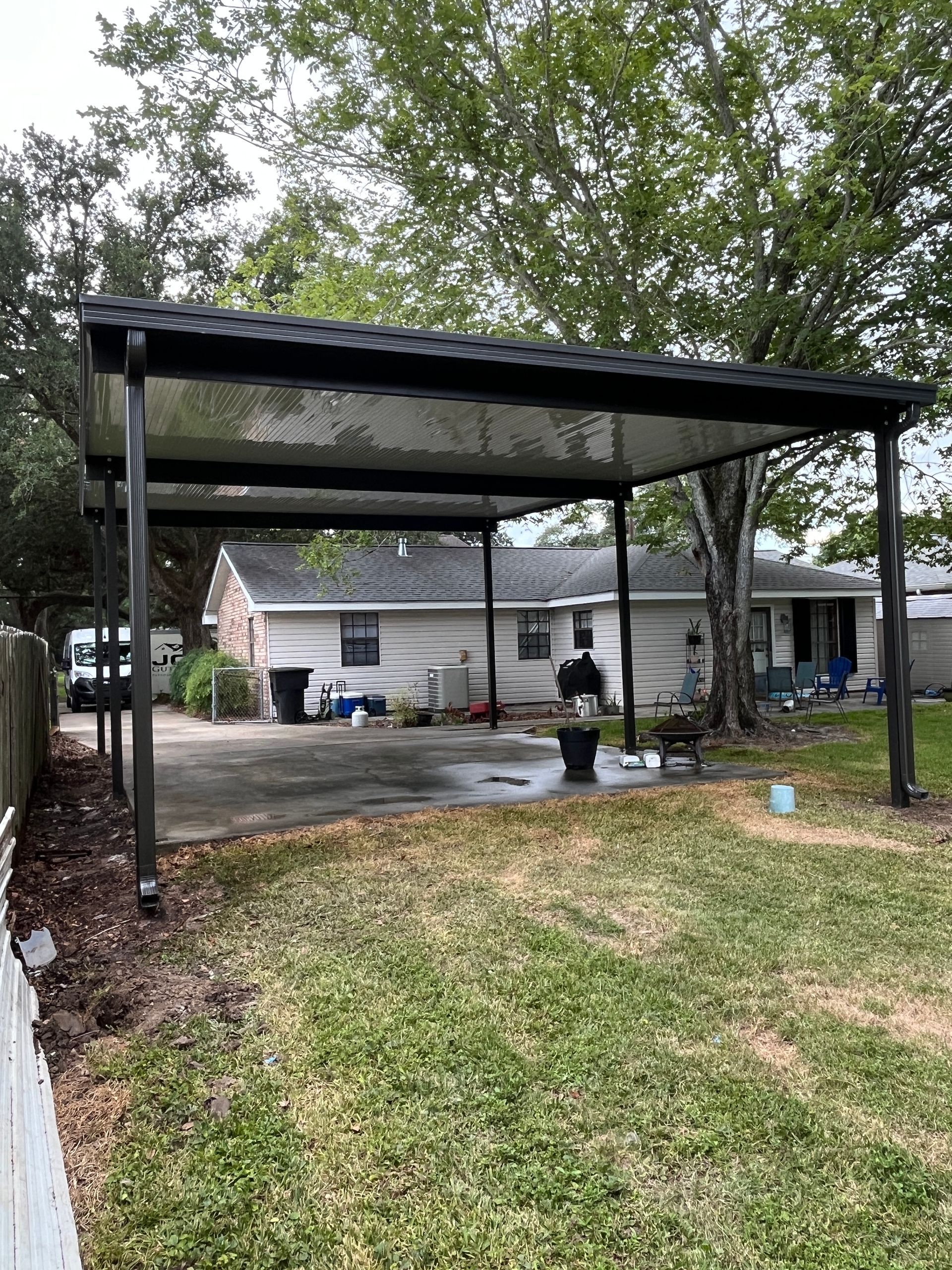A black metal carport stands over a concrete driveway next to a grassy yard and a single-story house.
