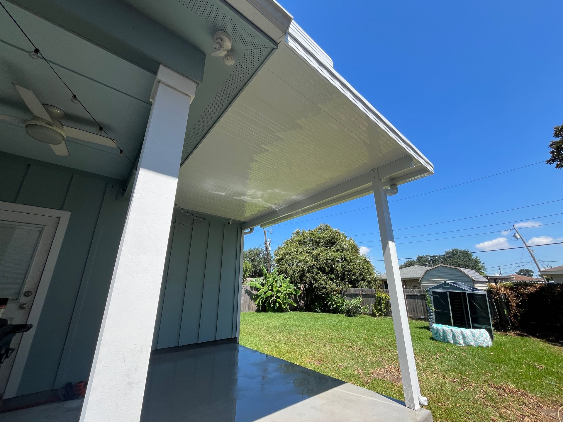 A view from a shaded, light-blue porch looking out at a grassy backyard under a clear blue sky on a sunny day.