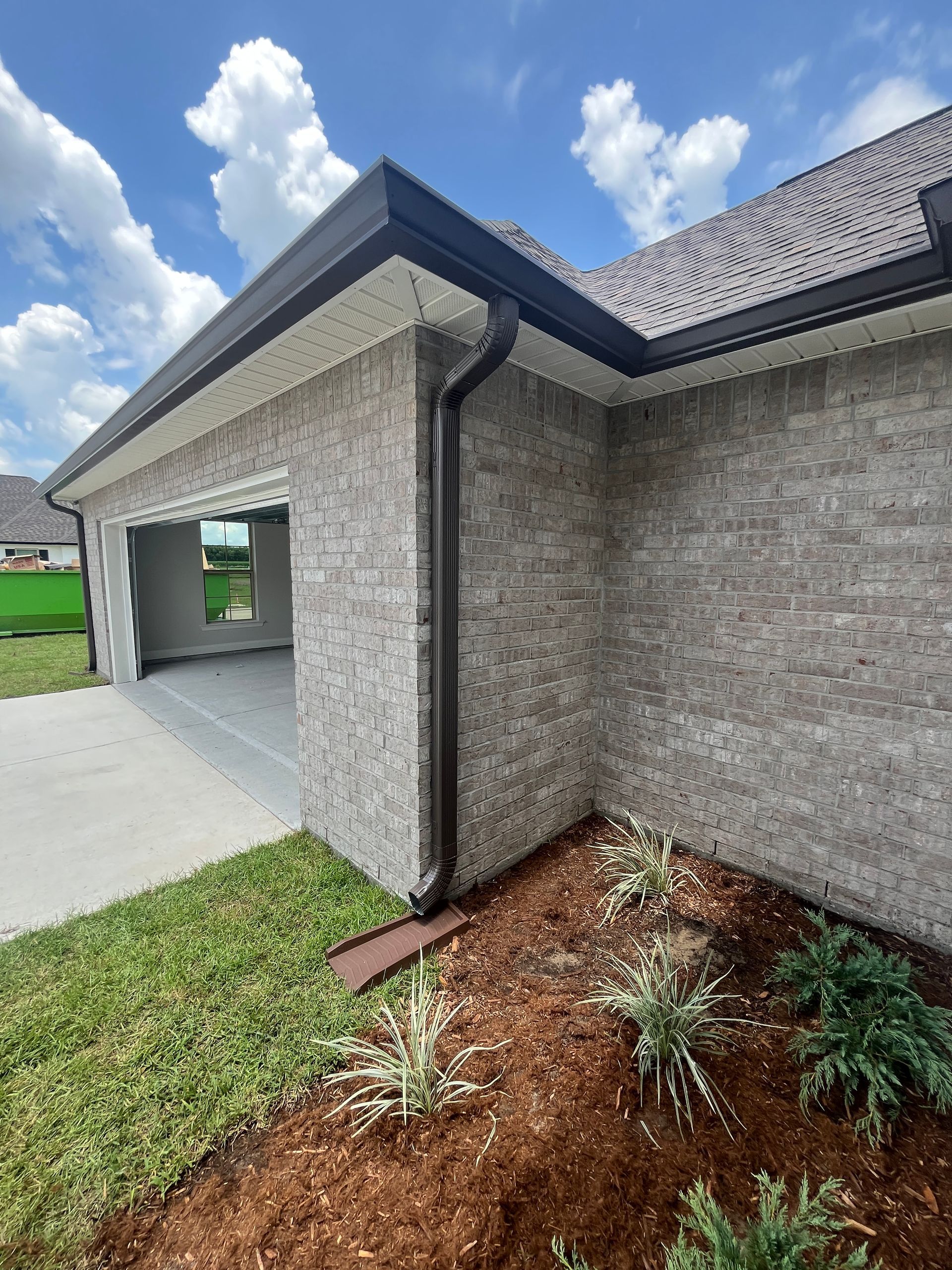 A low-angle view of a grey brick house corner with a black gutter, downspout, and landscaping with mulch.