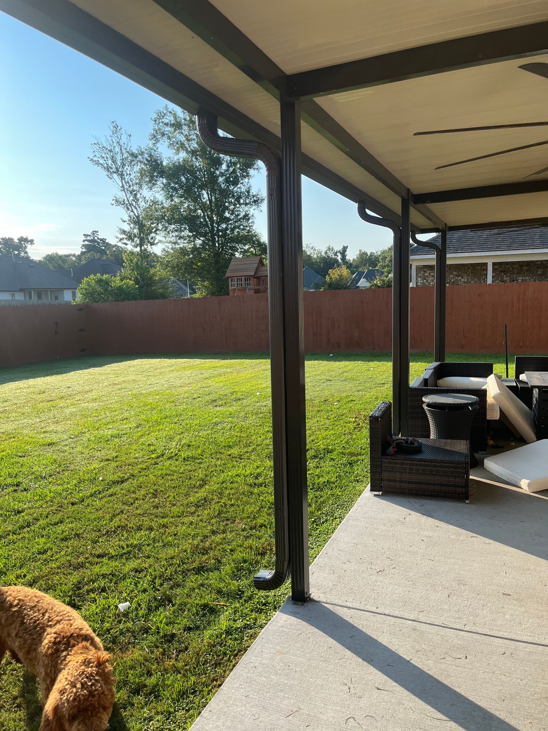 A patio with a brown support post and downspout, looking out over a grassy backyard with a brown fence and a dog.