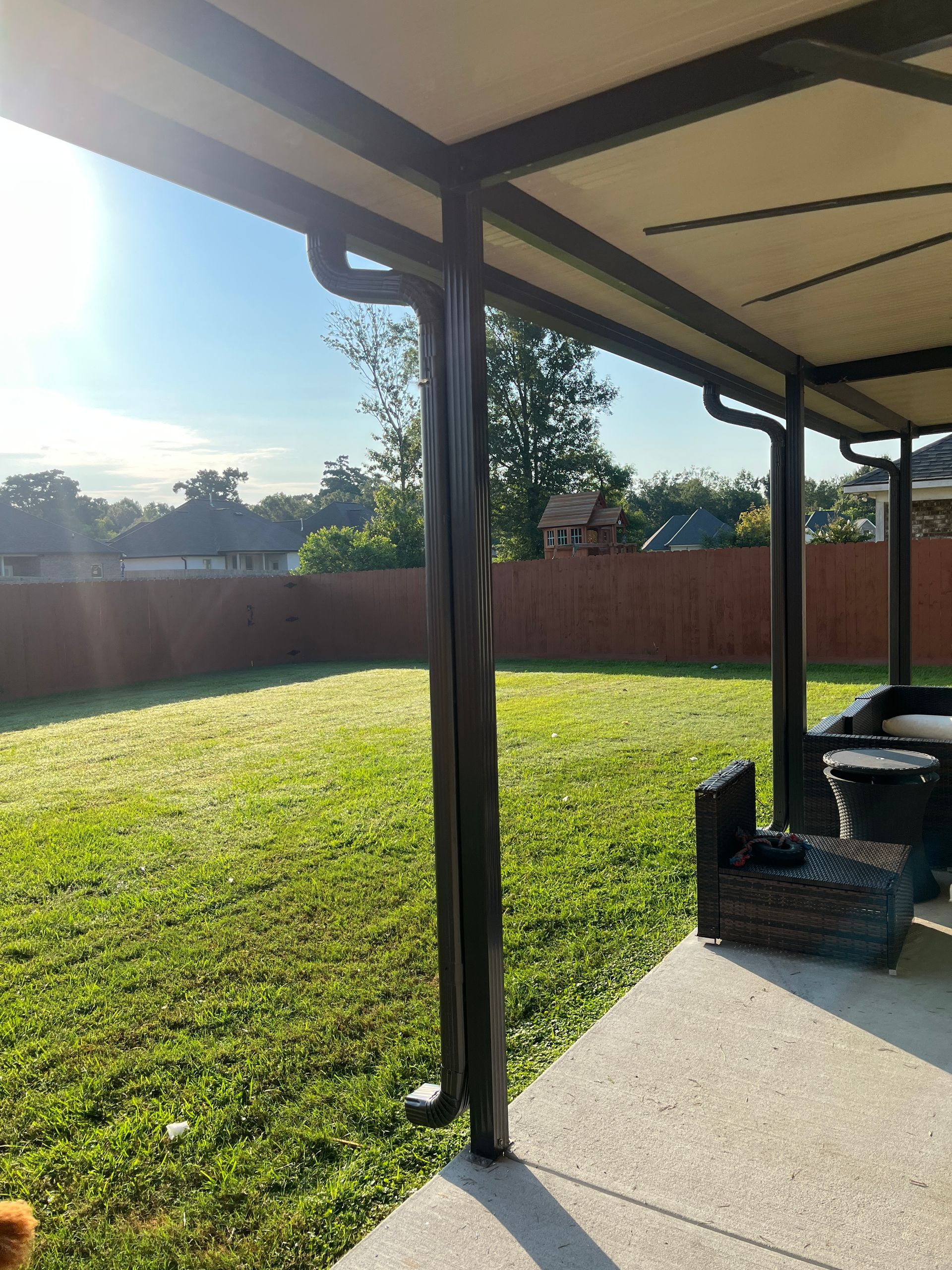 A sunny, grassy backyard viewed from a shaded concrete patio, featuring a brown fence and a nearby outdoor light fixture.