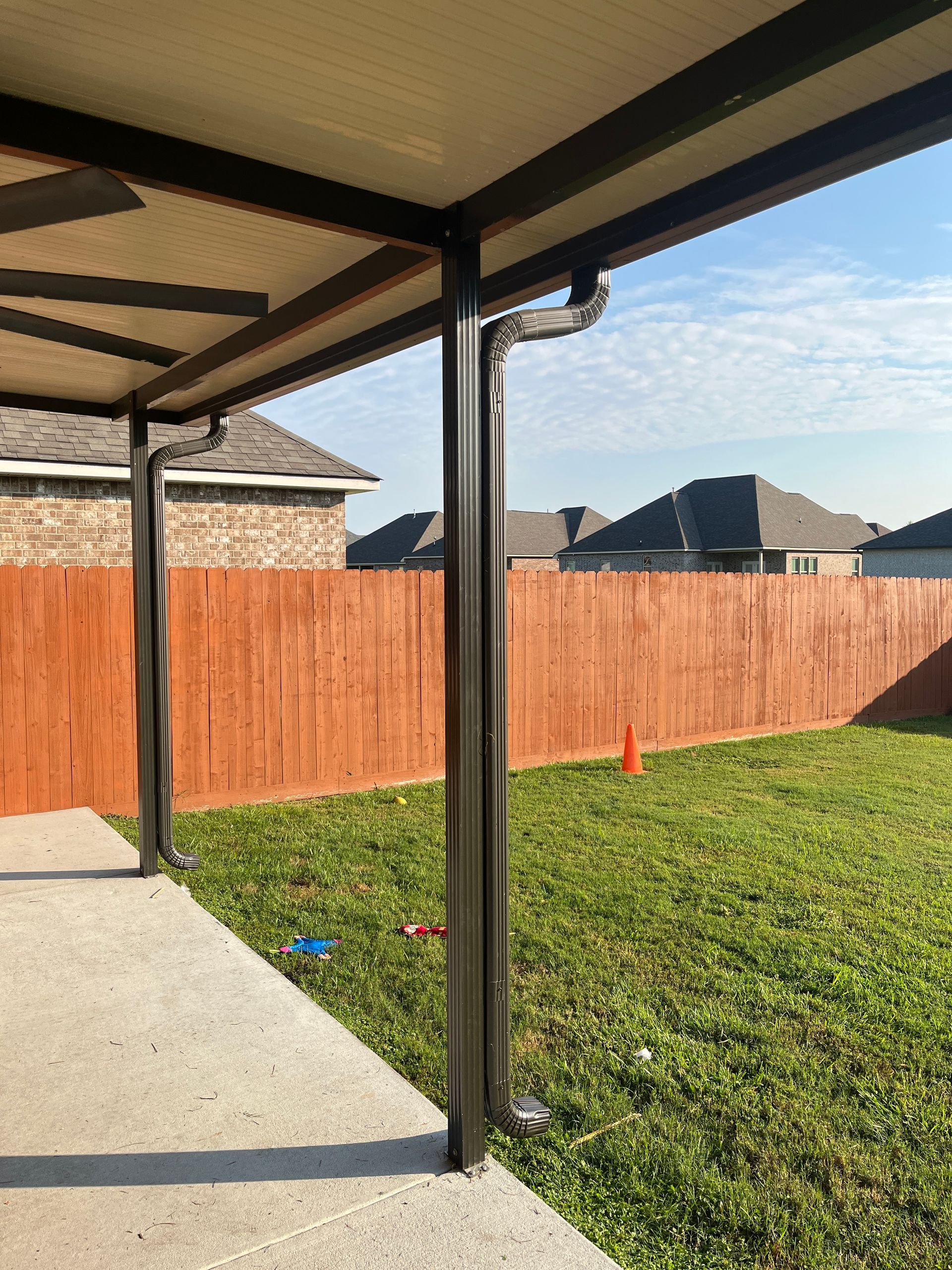 A black patio cover support column and gutter system in a backyard, with a wooden fence and houses in the background.