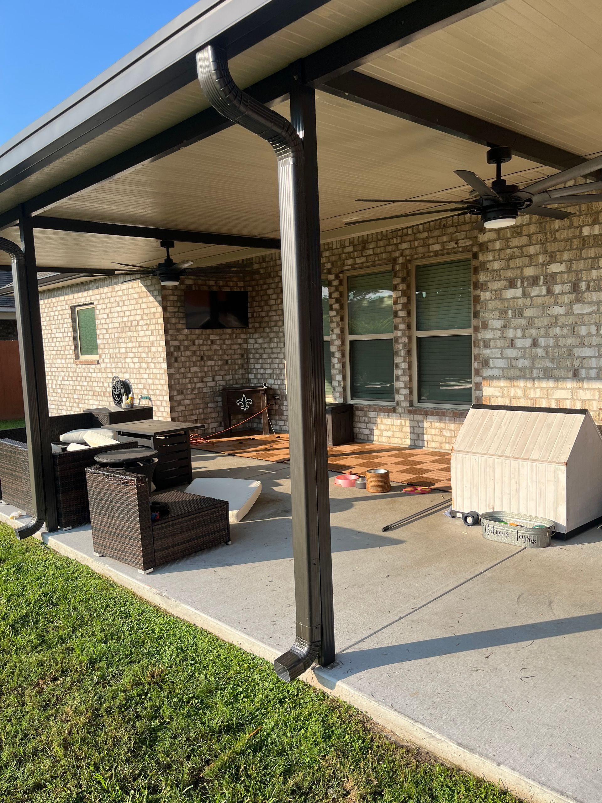 A concrete patio covered by a dark metal roof, featuring outdoor furniture, a television, and a dog house.