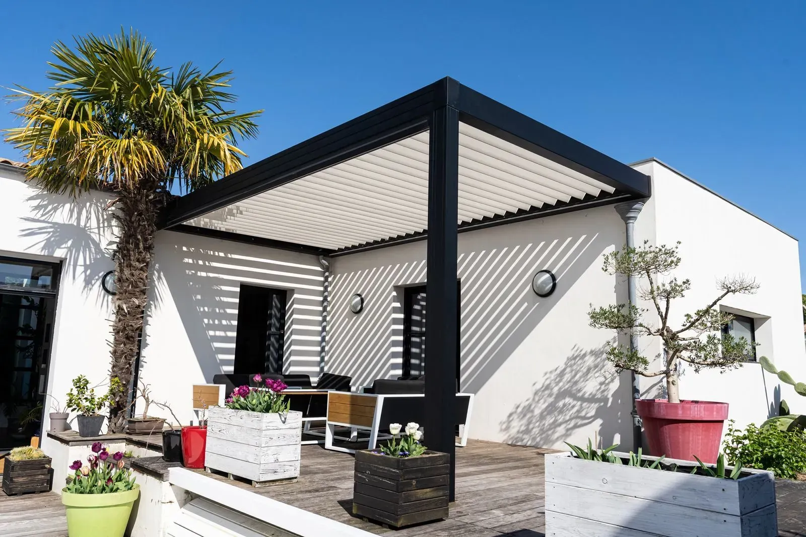 Modern white house patio with black pergola, potted plants, and outdoor seating in bright sunlight