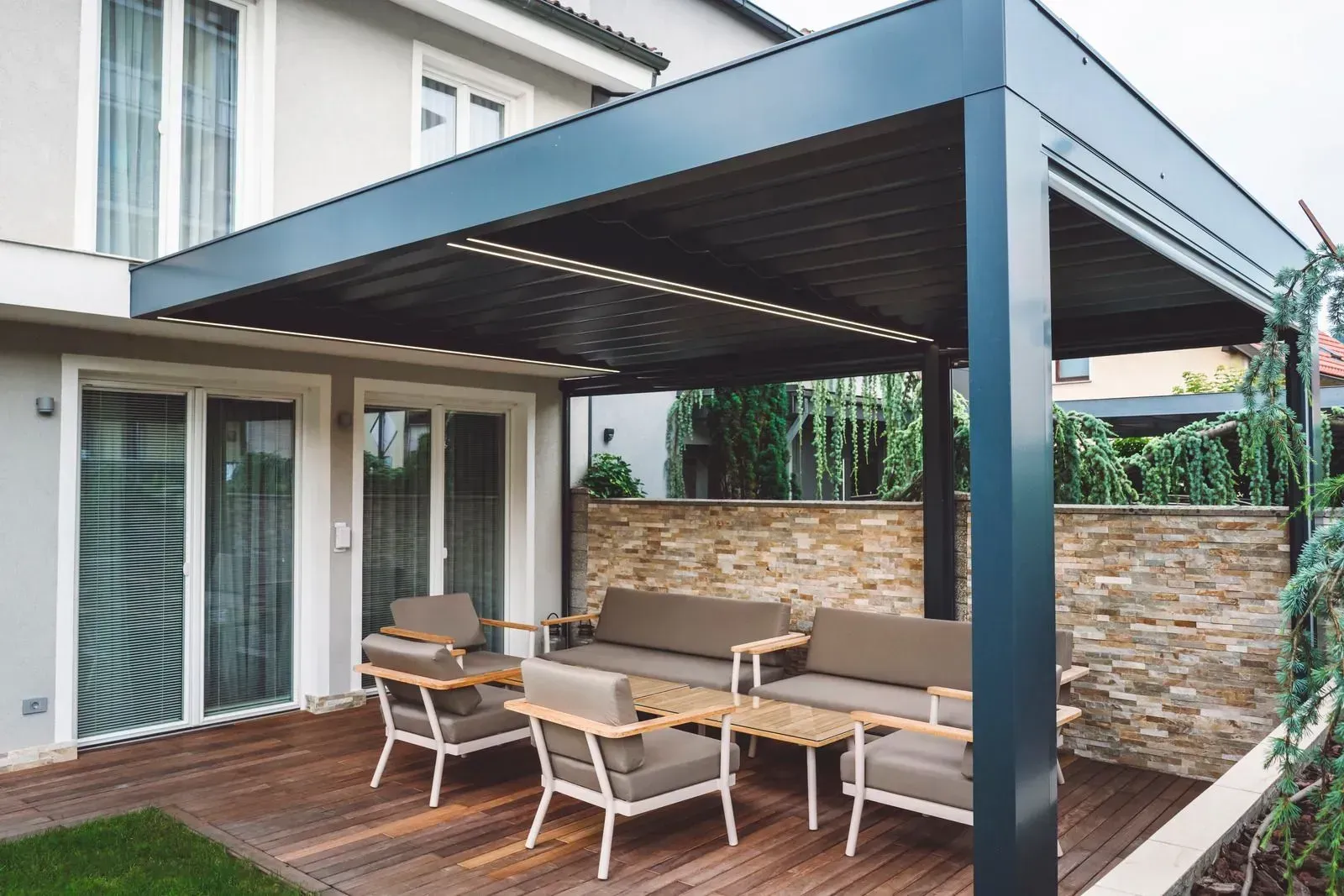 Covered patio with outdoor sofa and dining set beside a house, featuring a wood deck and stone wall