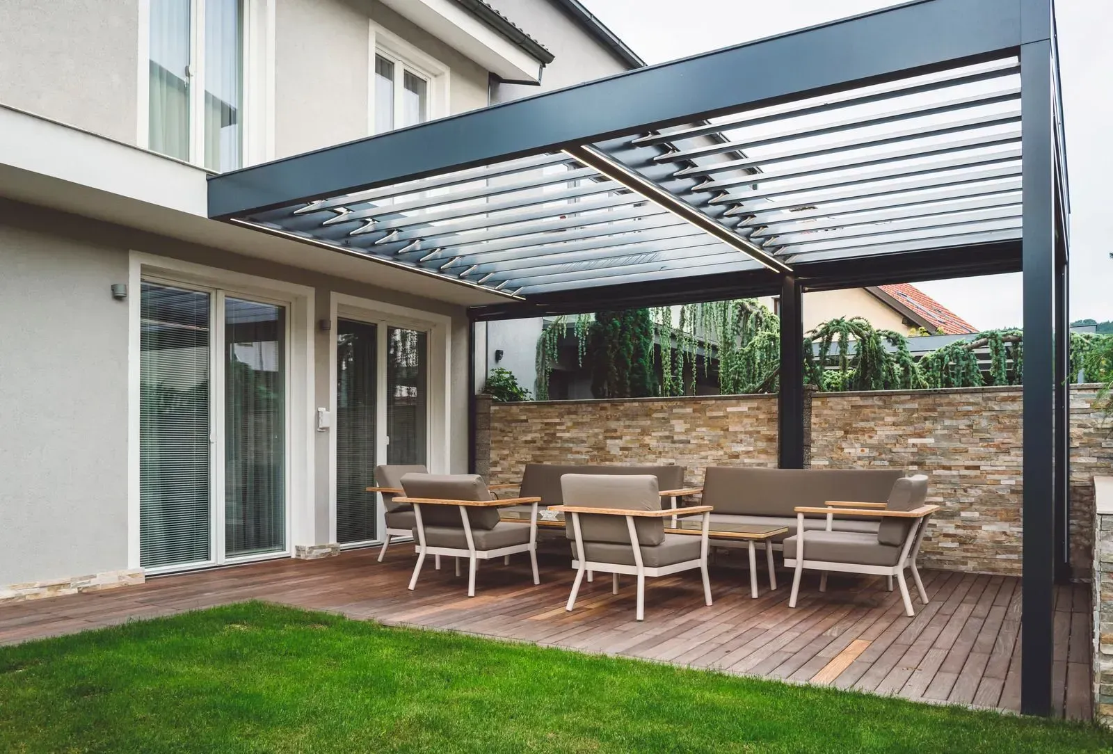 Modern patio with wooden deck, outdoor table and chairs, under a gray pergola beside a house
