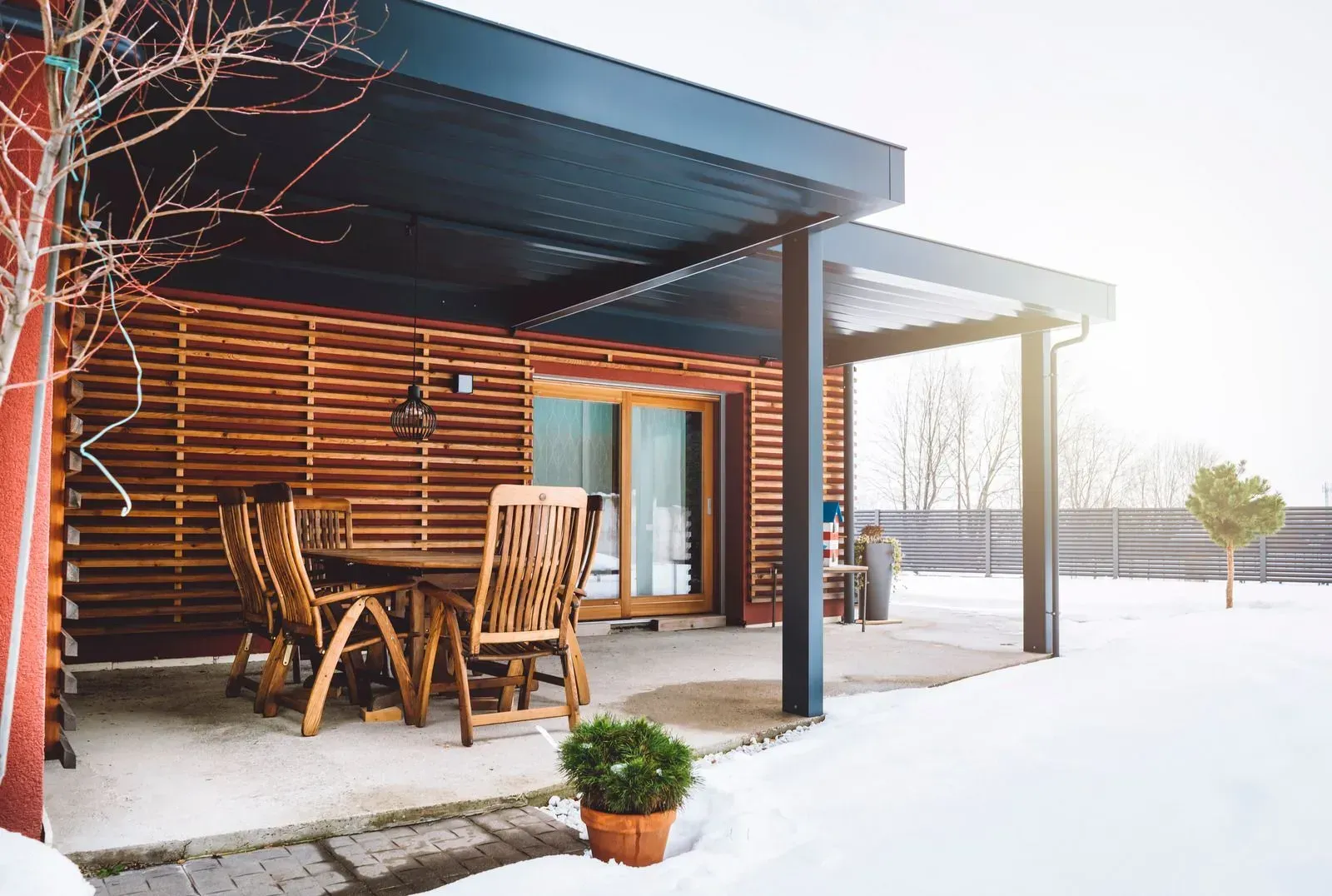 Snowy cabin patio with wooden chairs, table, and potted plant in bright sunlight