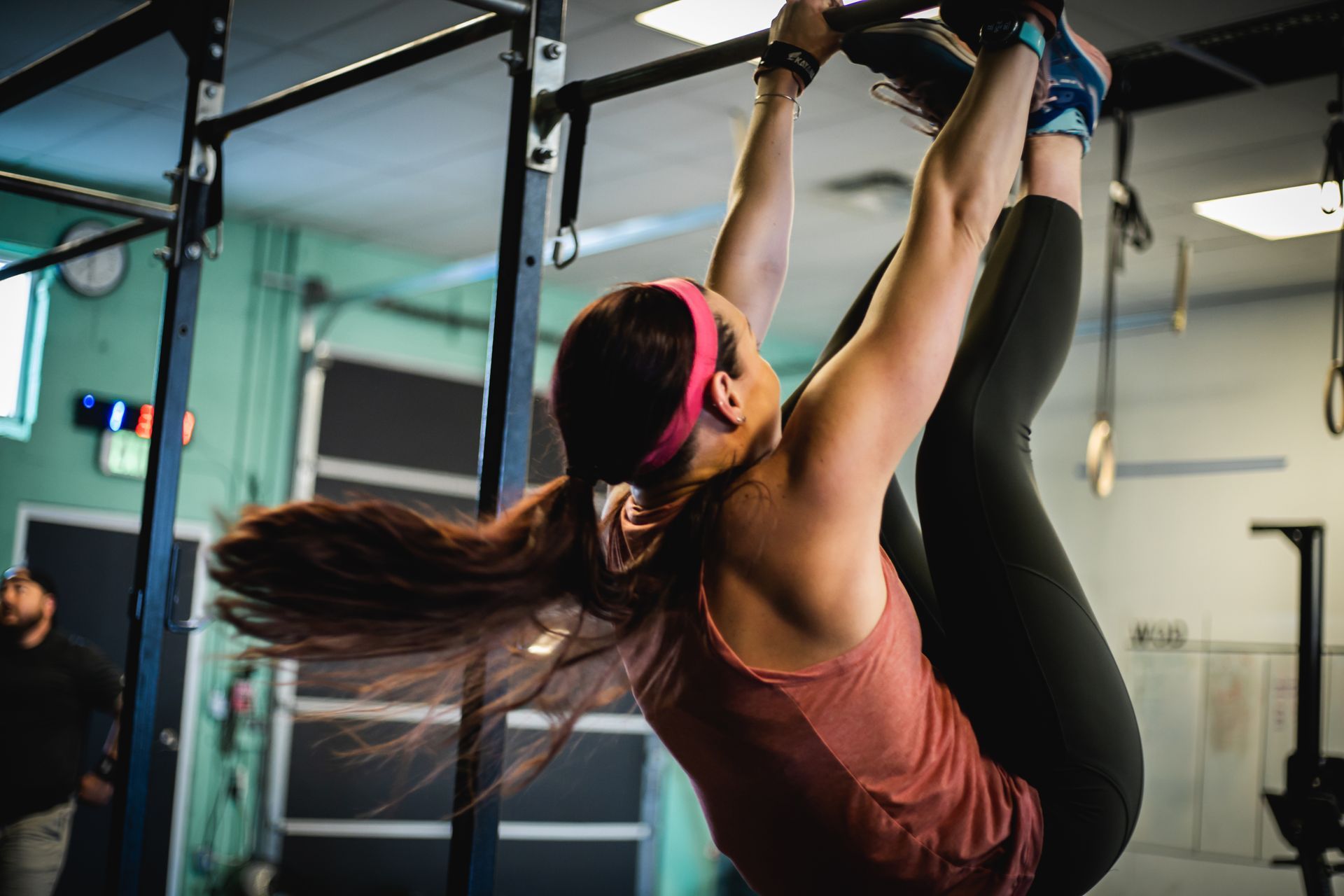 Woman doing a hanging leg raise in a CrossFit gym in Lakewood, CO, wearing a coral tank top and black leggings.